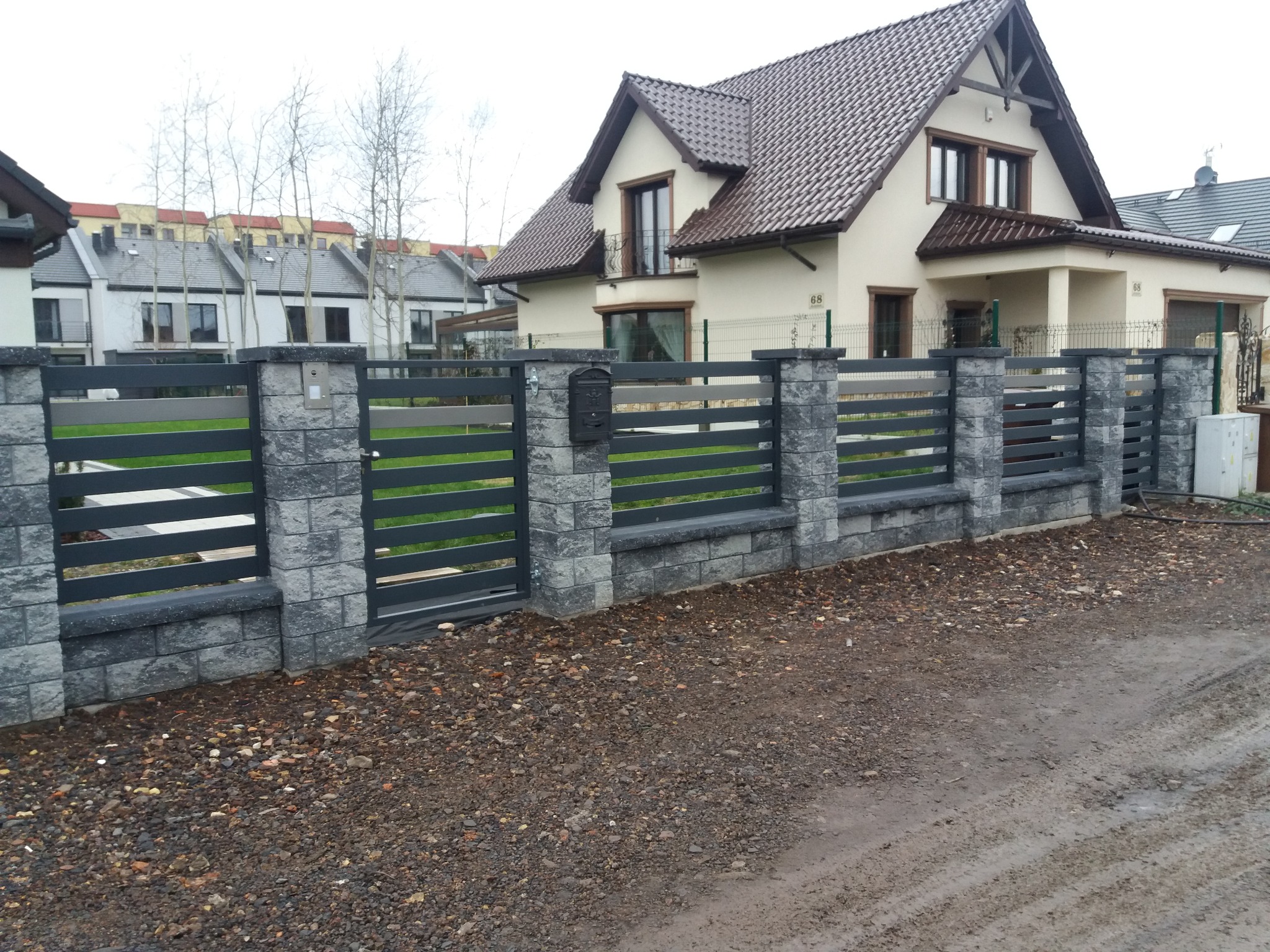 Nowoczesne ogrodzenie panelowe z elementami imitującymi kamień, szare panele, skrzynka pocztowa, dom jednorodzinny w tle.