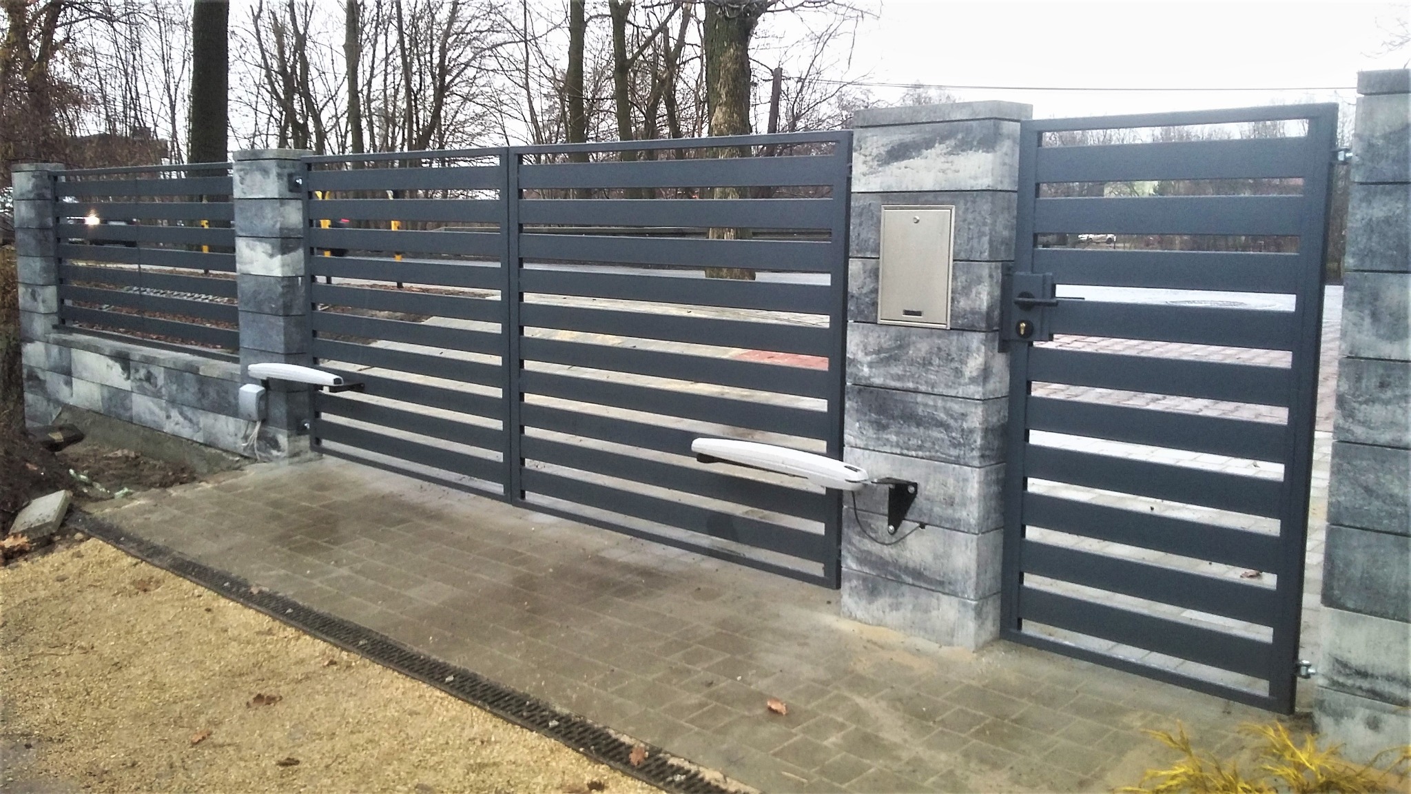 Nowoczesna, pozioma, szara brama wjazdowa i furtka z automatycznym napędem, słupki z szarej kostki, skrzynka na listy, widok z zewnątrz.
