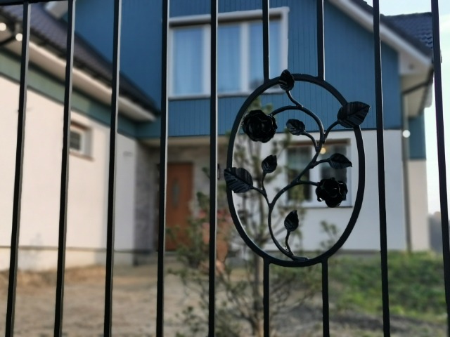 Kute, metalowe ogrodzenie z dekoracyjnym elementem w kształcie owalu z motywem roślinnym róż, na tle domu z niebieską elewacją.