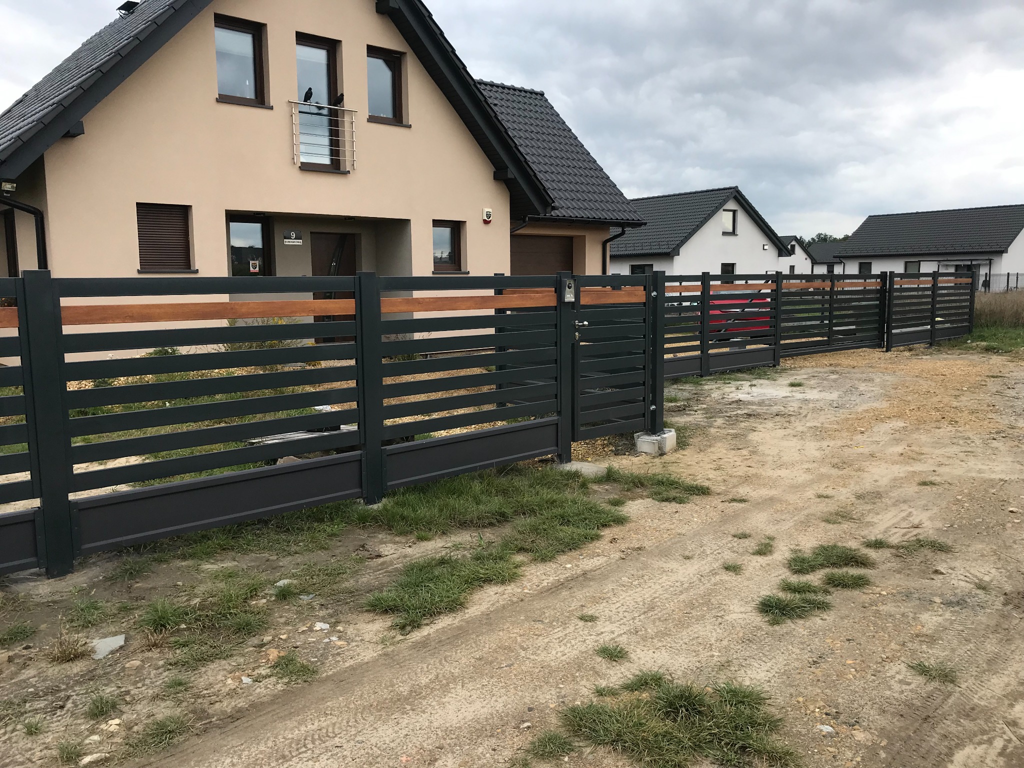 Nowoczesne ogrodzenie panelowe z elementami drewnianymi, szara brama wjazdowa, dom jednorodzinny w tle, pochmurne niebo.