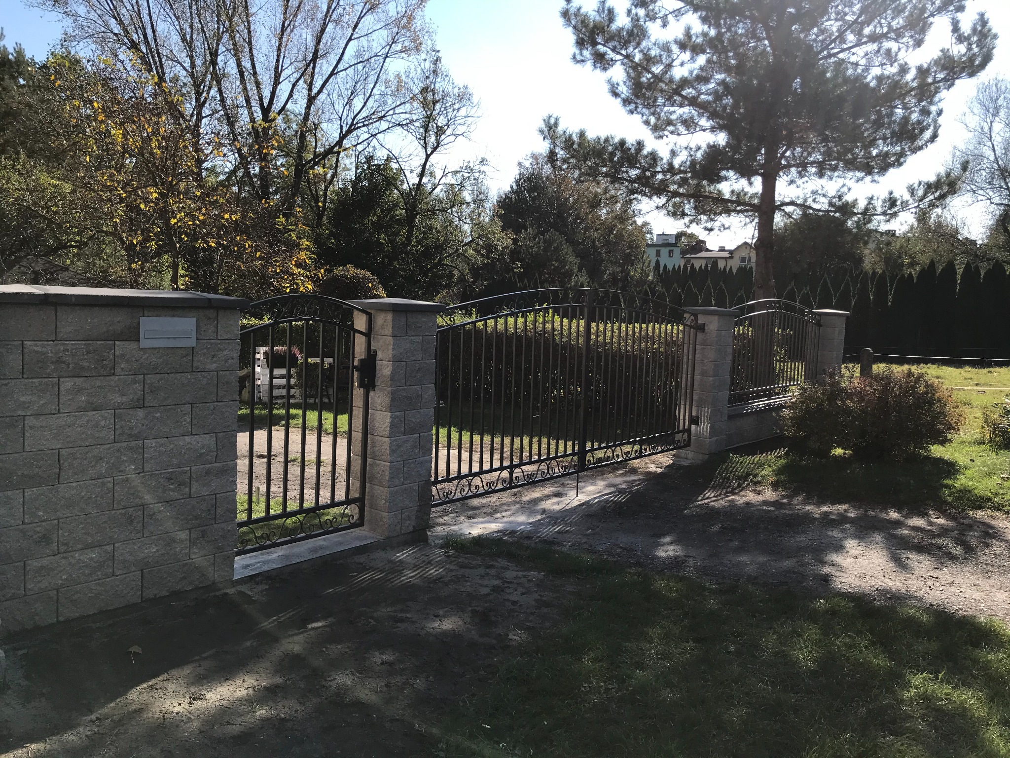 Czarna, metalowa brama wjazdowa z dekoracyjnymi elementami, osadzona na słupkach z szarej kostki, widok na posesję z zielenią i drzewami.