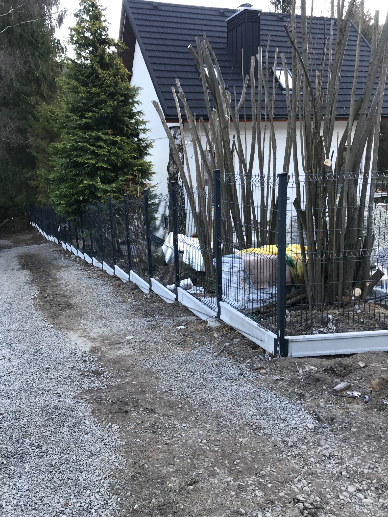 Nowo zamontowane, panelowe ogrodzenie z podmurówką, widok z perspektywy ścieżki z szutru, w tle dom z ciemnym dachem i przycięte krzewy.