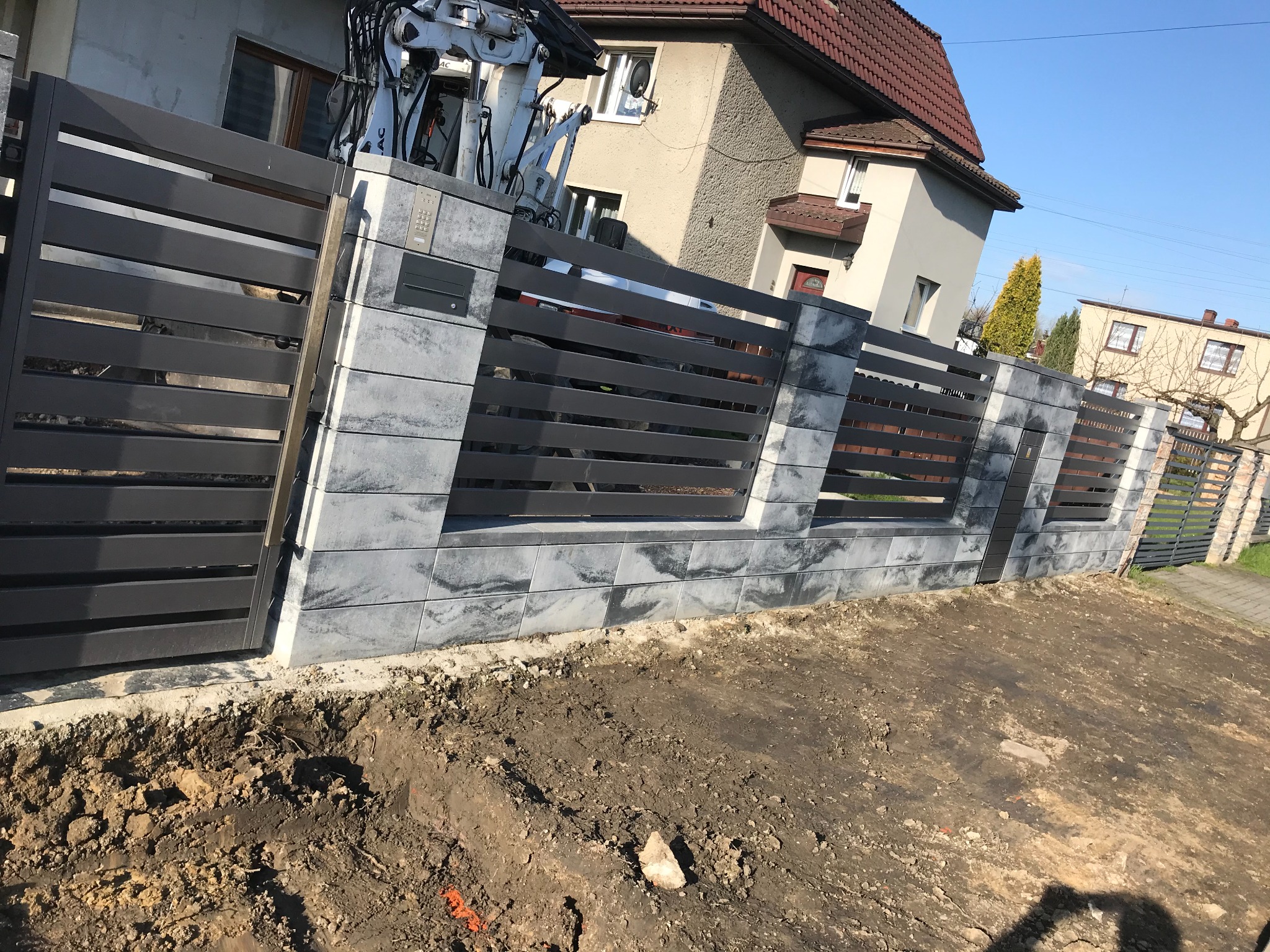 Nowoczesne ogrodzenie panelowe z elementami betonowymi w trakcie montażu; widoczny dźwig, niezagospodarowany teren.