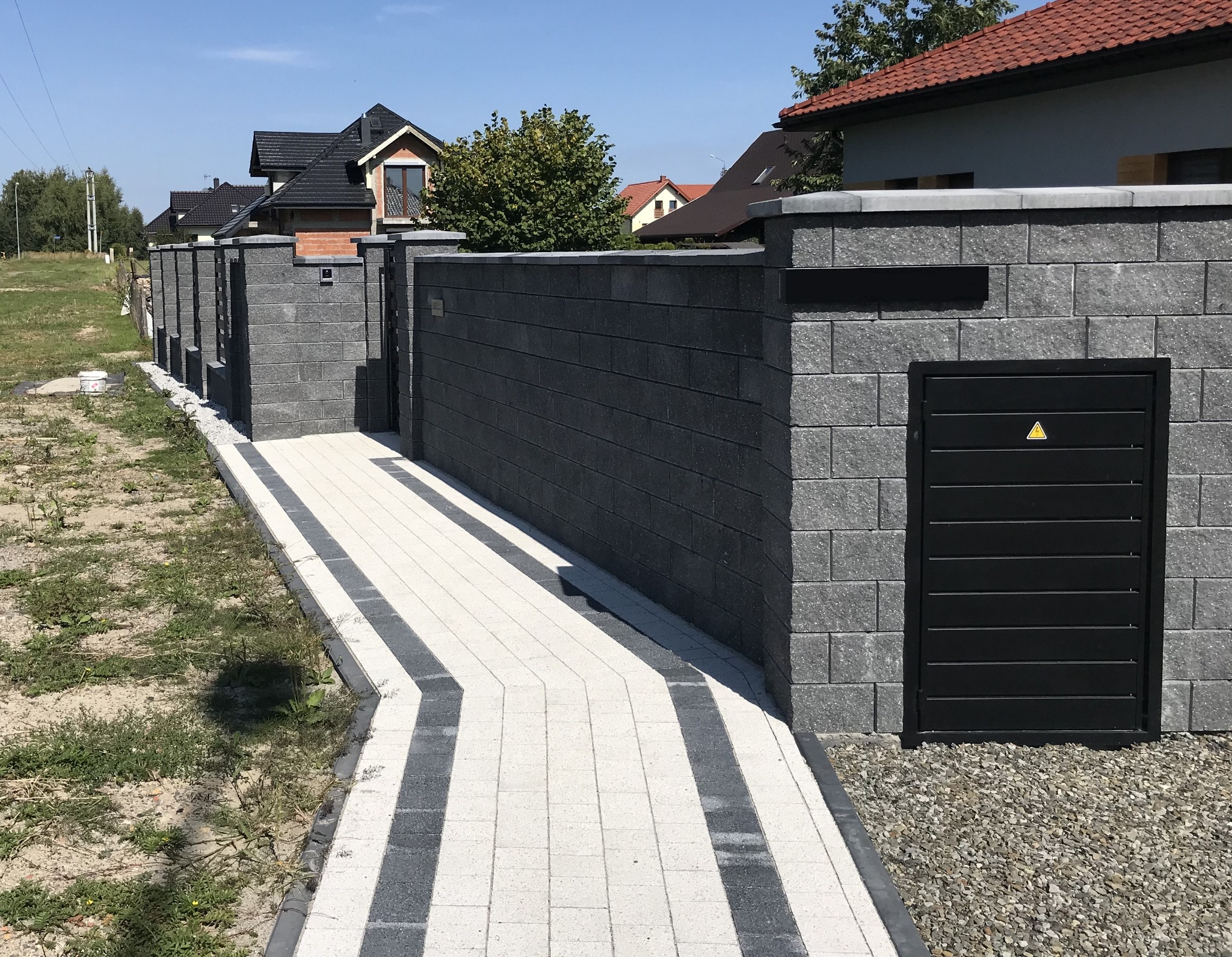 Nowoczesne ogrodzenie z szarej kostki betonowej z czarnymi akcentami: skrzynka na listy i drzwiczki rewizyjne z żółtym symbolem ostrzegawczym.