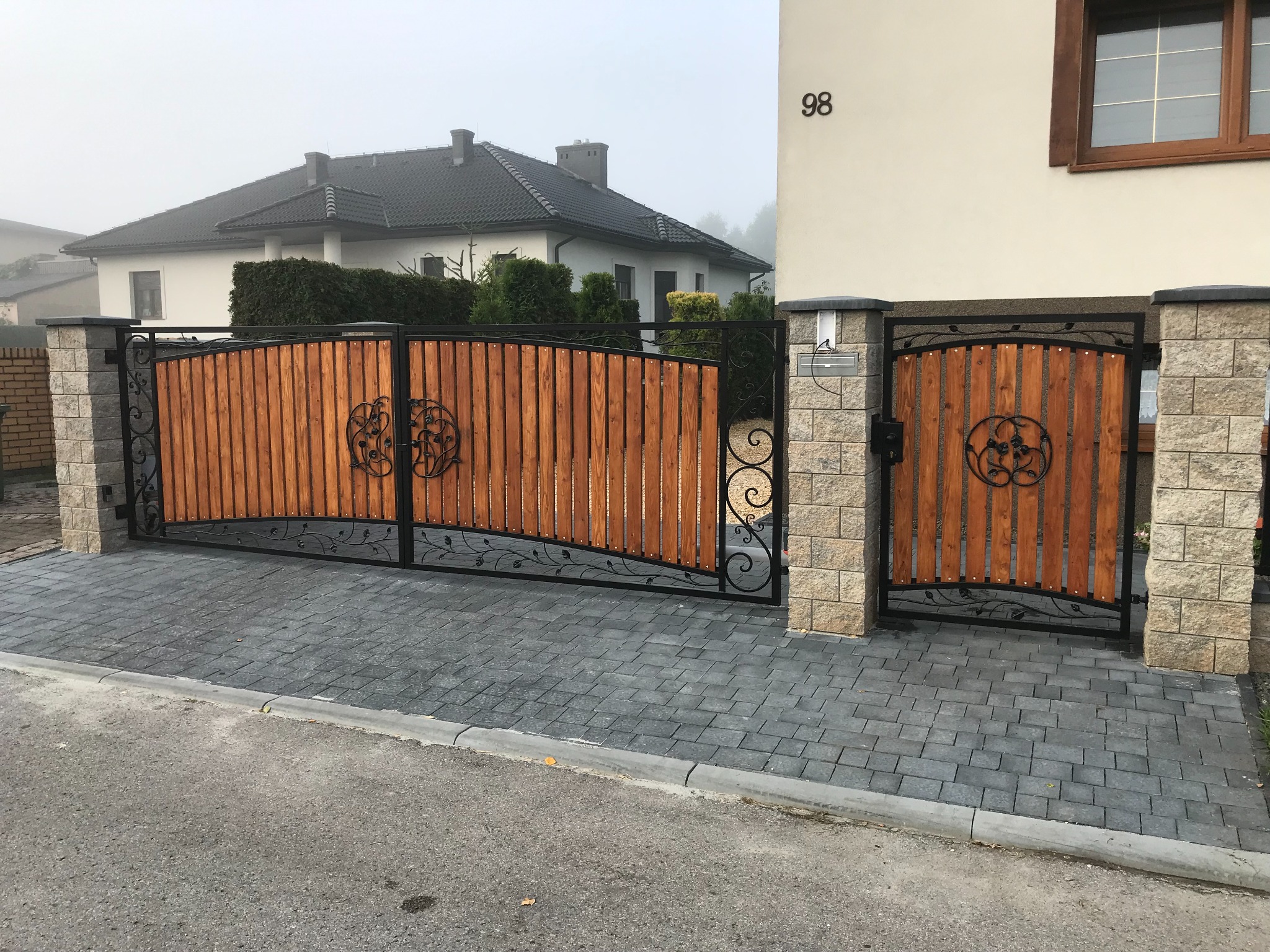Nowoczesna brama wjazdowa i furtka z drewnianym wypełnieniem i ozdobnymi elementami z kutego żelaza, na tle domu z numerem 98, w porannym, mglistym świetle.