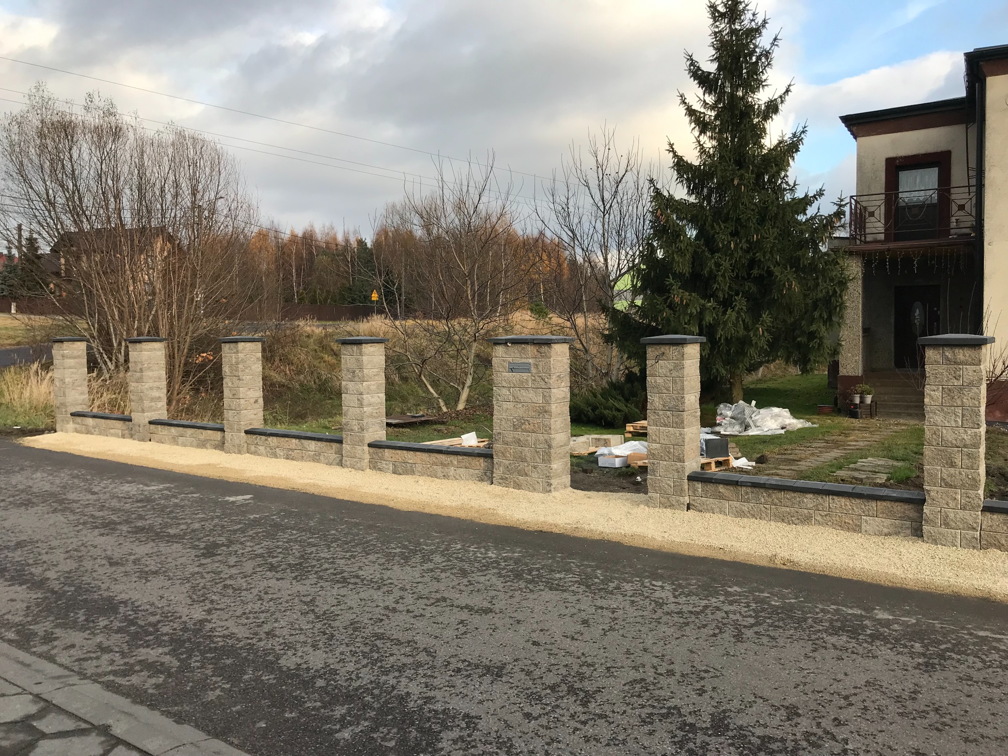 Niski, modułowy płot z szarego kamienia łupanego z czarnymi nakładkami, oddzielający posesję od asfaltowej drogi, z widocznymi materiałami budowlanymi na trawniku za płotem.