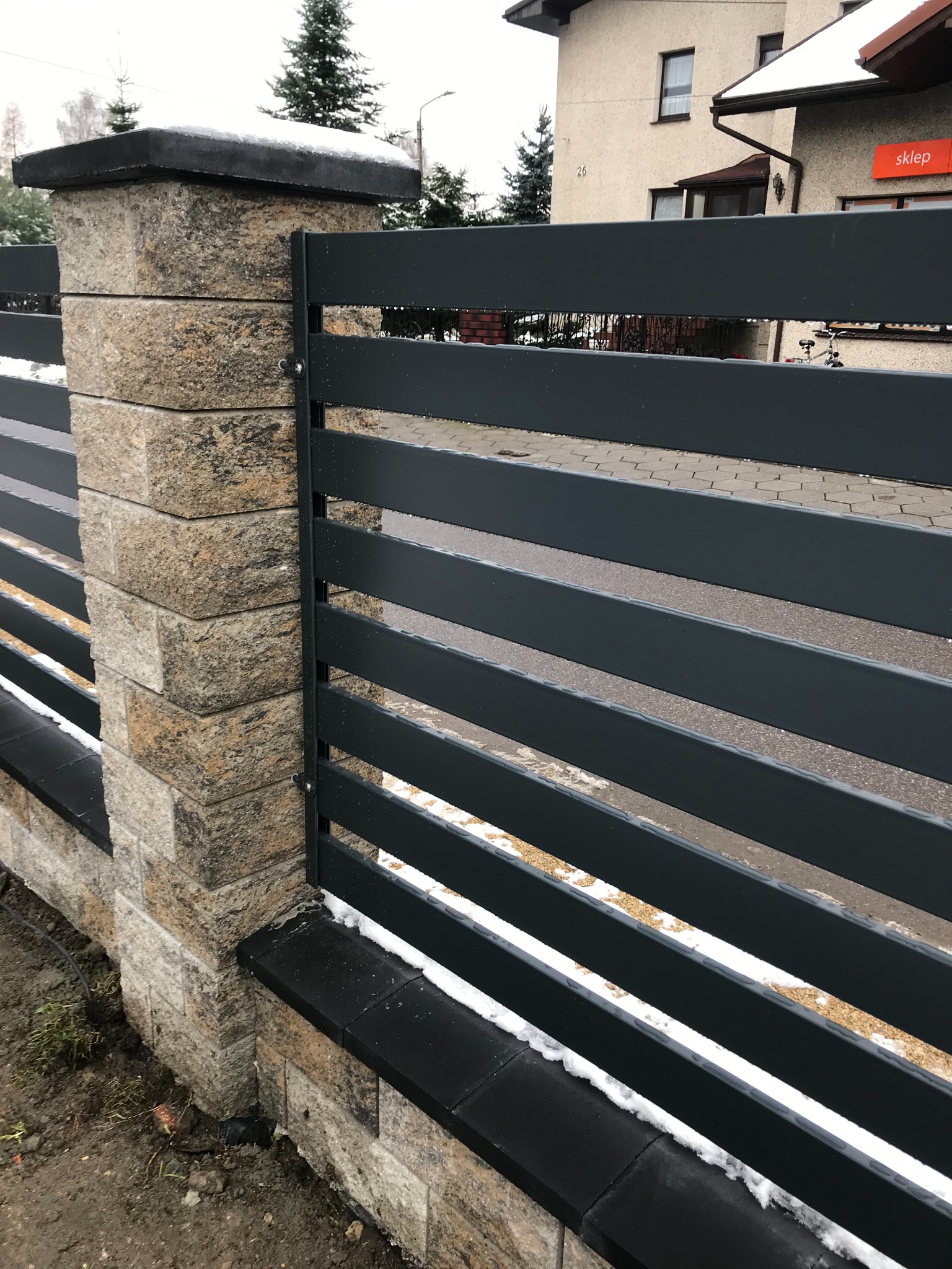 Nowoczesne ogrodzenie panelowe z grafitowymi przęsłami i słupkami z kamienia naturalnego, lekko przyprószone śniegiem, widok z bliska na fragment ogrodzenia.