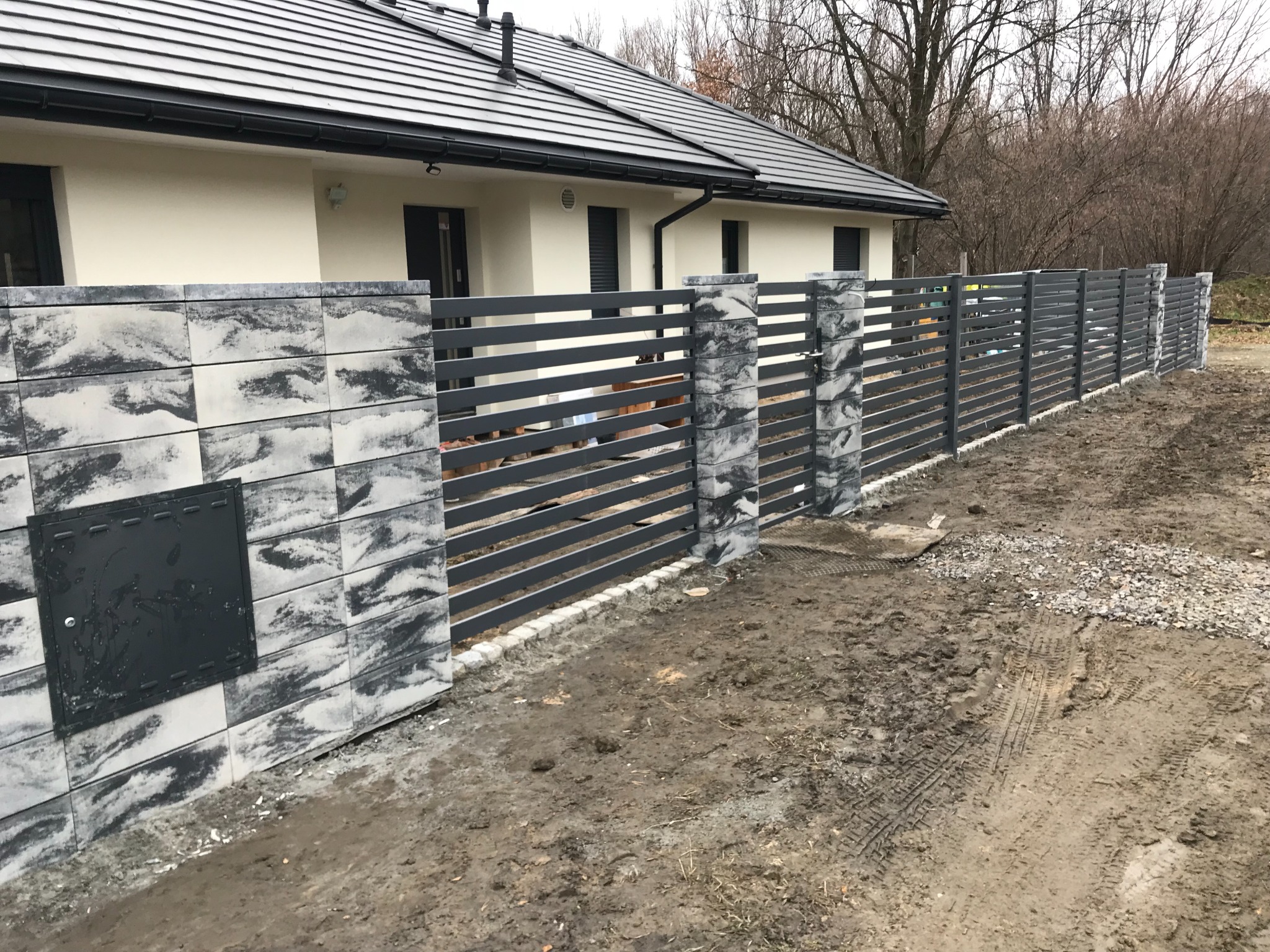 Nowoczesne ogrodzenie panelowe z podmurówką z szarych bloczków imitujących kamień, zamontowane przed domem z ciemną dachówką, widoczne ślady opon na błotnistym terenie.