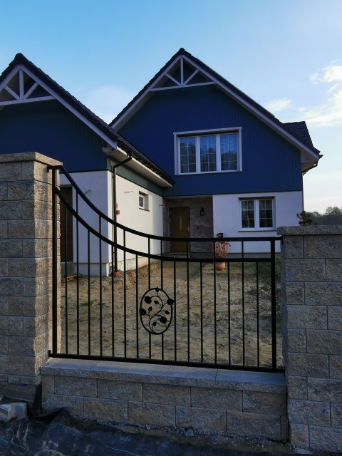 Czarna brama wjazdowa z ozdobnym motywem roślinnym, osadzona na kamiennych słupkach i podmurówce, na tle domu z niebieską elewacją i jasną fasadą.