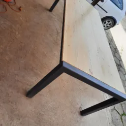 Stół z jasnego drewna z czarną, metalową ramą, widoczny na zewnątrz, obok zaparkowanego samochodu.