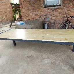 Stół z jasnego drewna z czarną, metalową ramą, ustawiony na betonowym podłożu przed ceglaną ścianą, z widocznym rowerem i jacuzzi w tle.