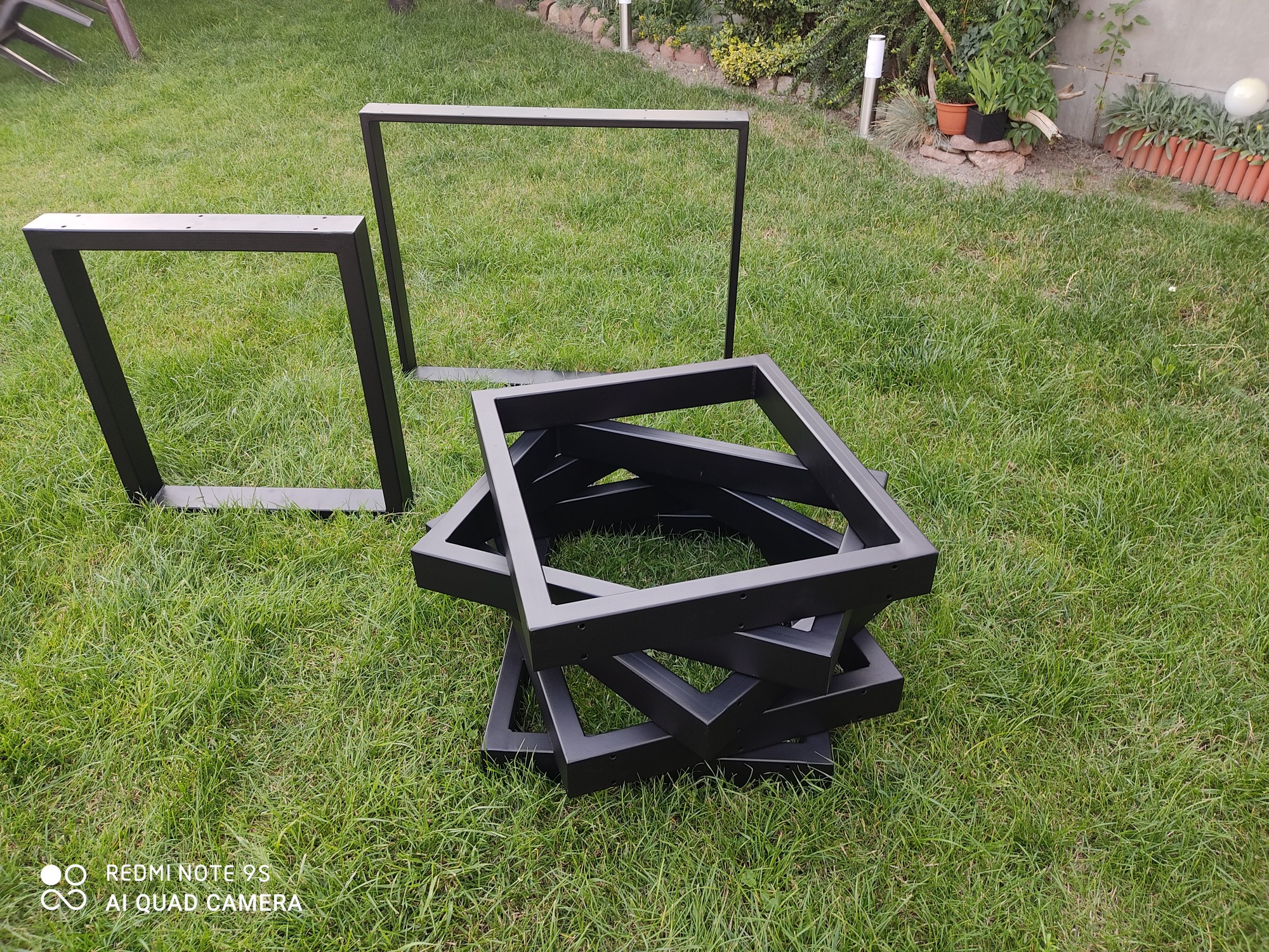 Czarne, metalowe kwadratowe ramy nóg stołowych, ułożone na trawie w ogrodzie, widoczne otwory montażowe.