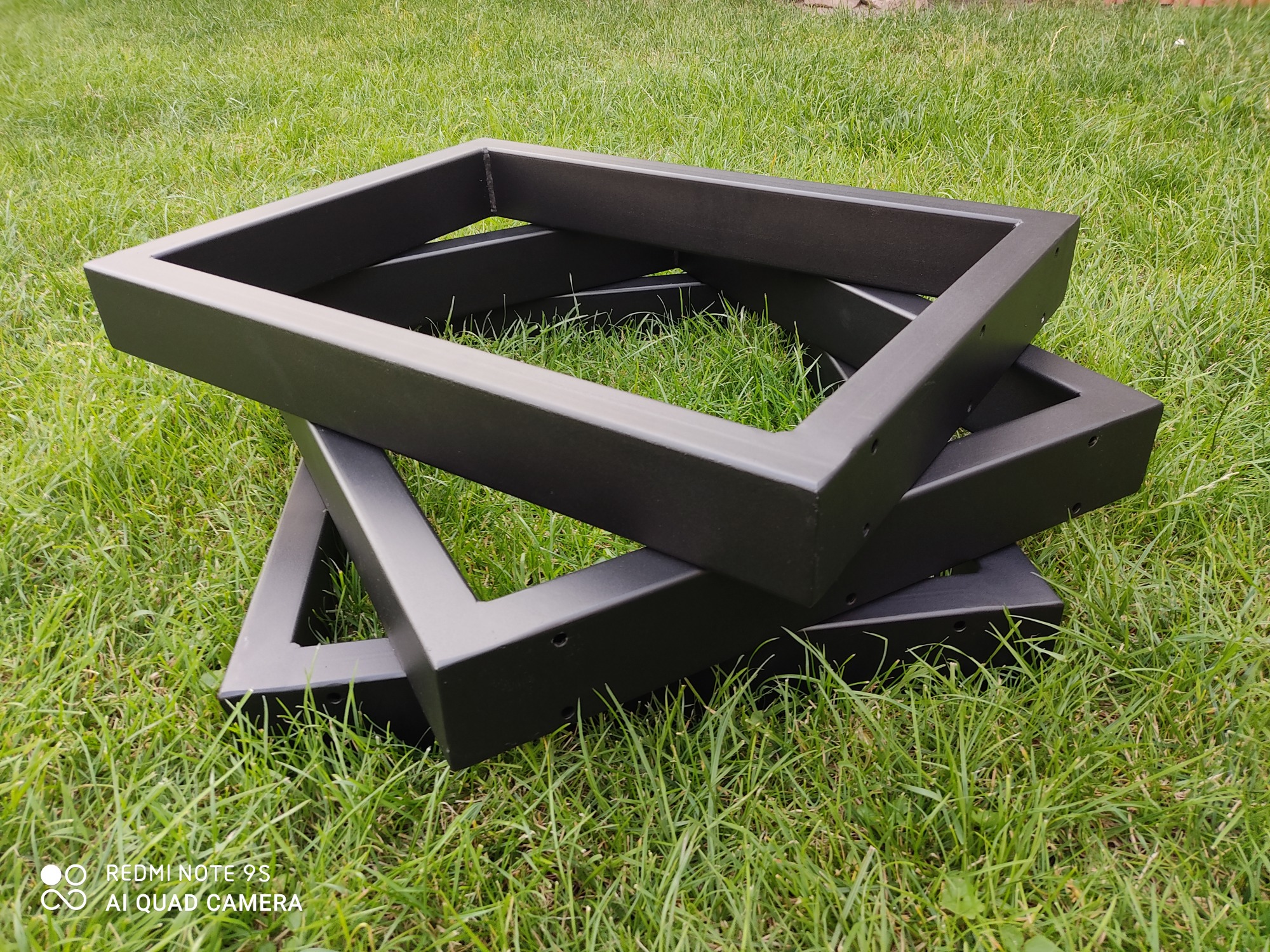 Stos trzech czarnych, spawanych, kwadratowych ram z metalowych profili na zielonej trawie. Widoczne spawy i otwory montażowe.