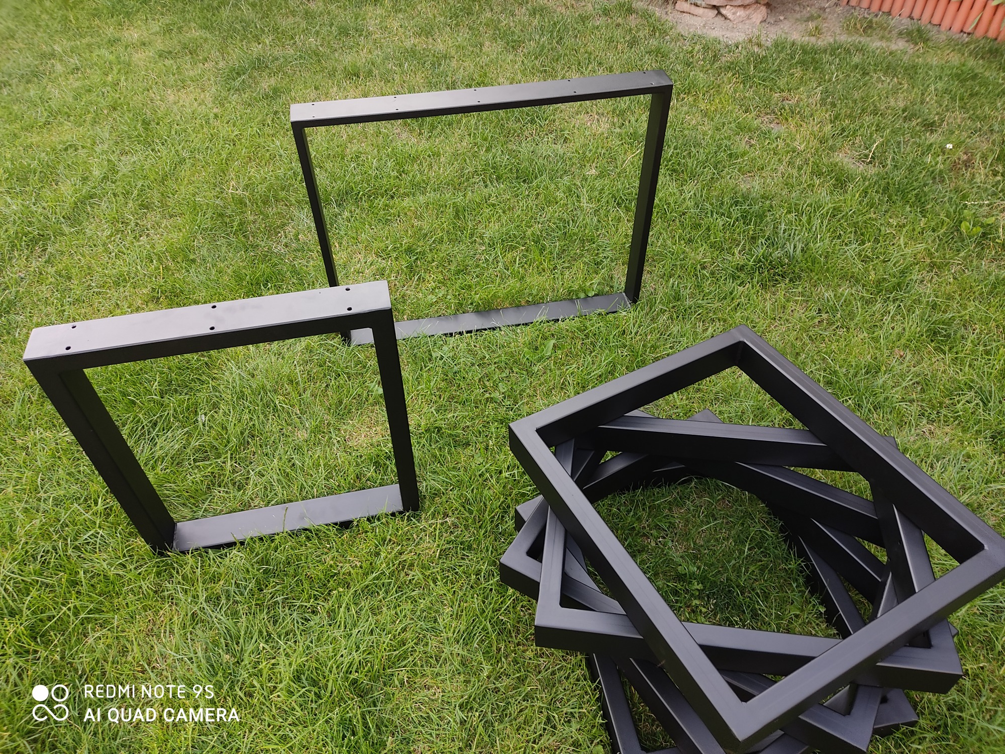 Czarne, metalowe ramy stołów, niektóre z otworami montażowymi, leżące na trawie w słoneczny dzień.