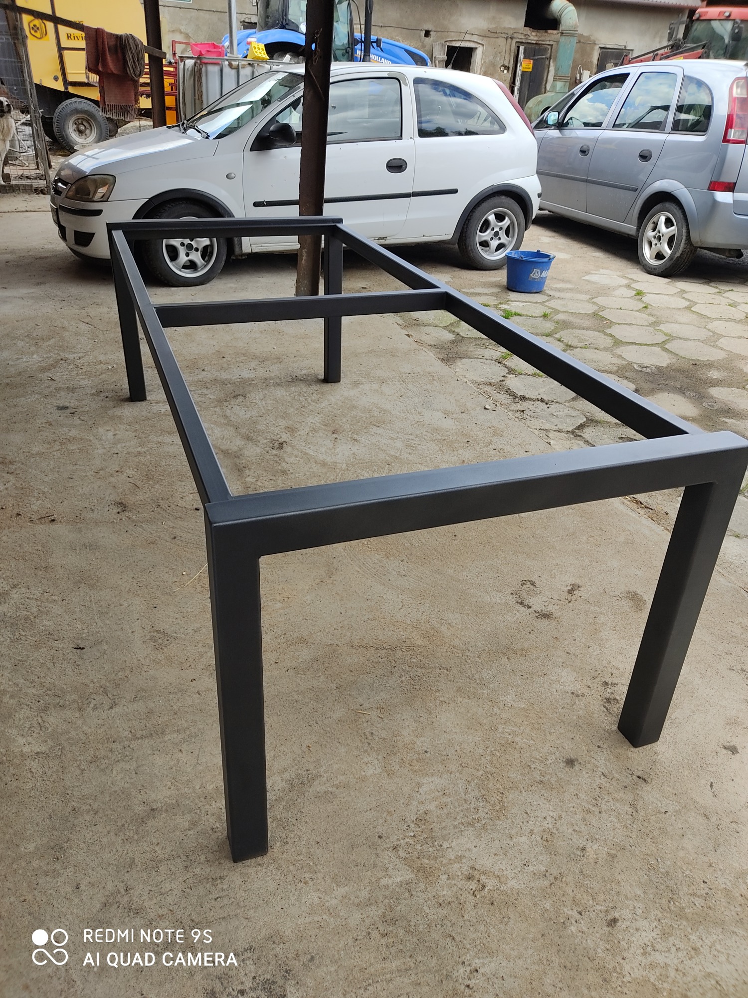 Stalowa rama stołu z profili zamkniętych, pomalowana proszkowo na czarno, na betonowym podłożu, z samochodami w tle.