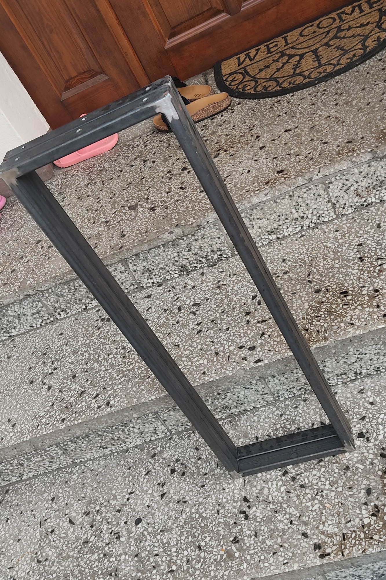 Spawana, prostokątna rama z metalowych profili na schodach o mozaikowej powierzchni, widoczne ślady spawania na rogach.