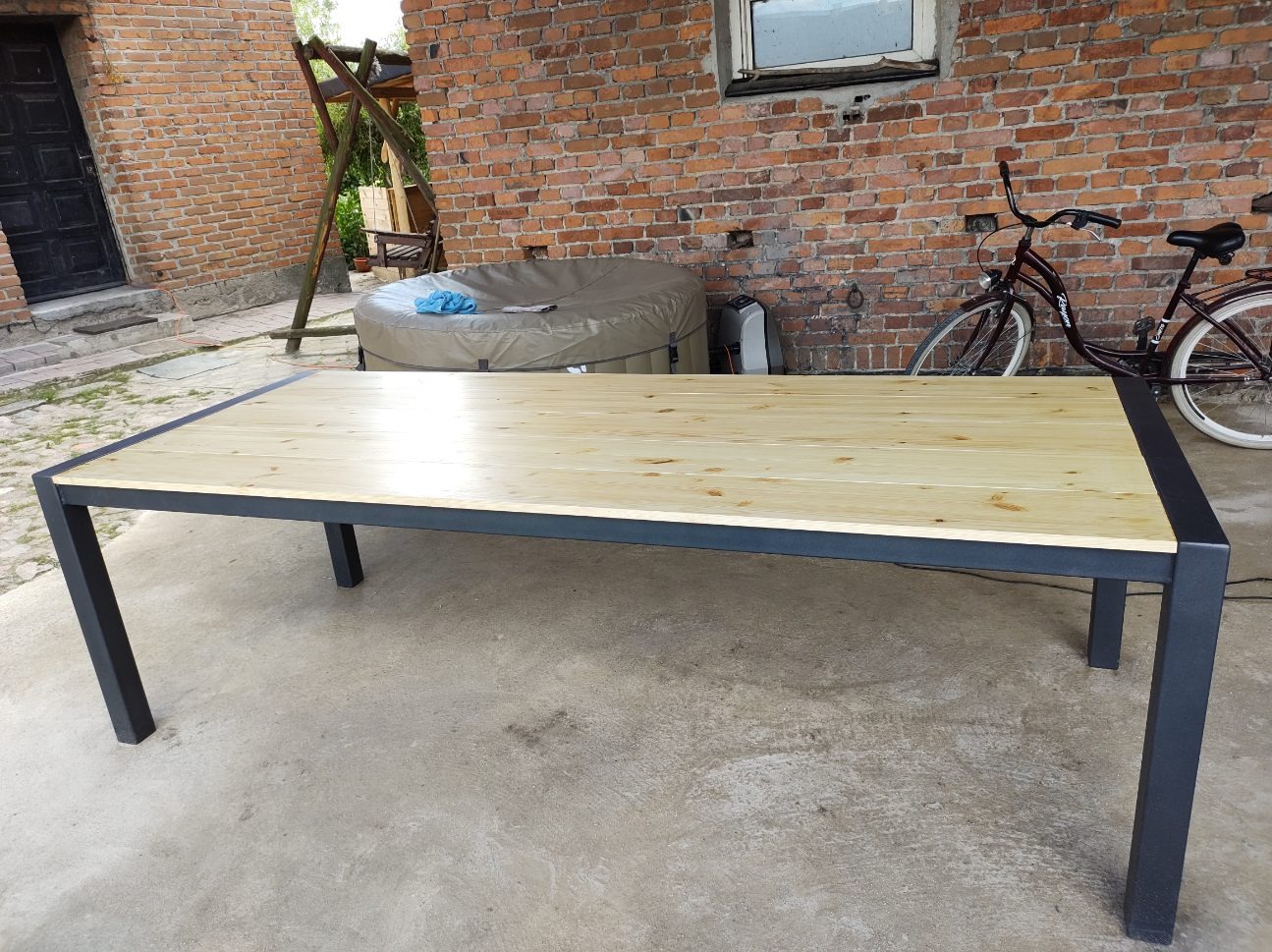 Stół z jasnego drewna z czarną, metalową ramą, ustawiony na betonowym podłożu przed ceglaną ścianą, z widocznym rowerem i jacuzzi w tle.