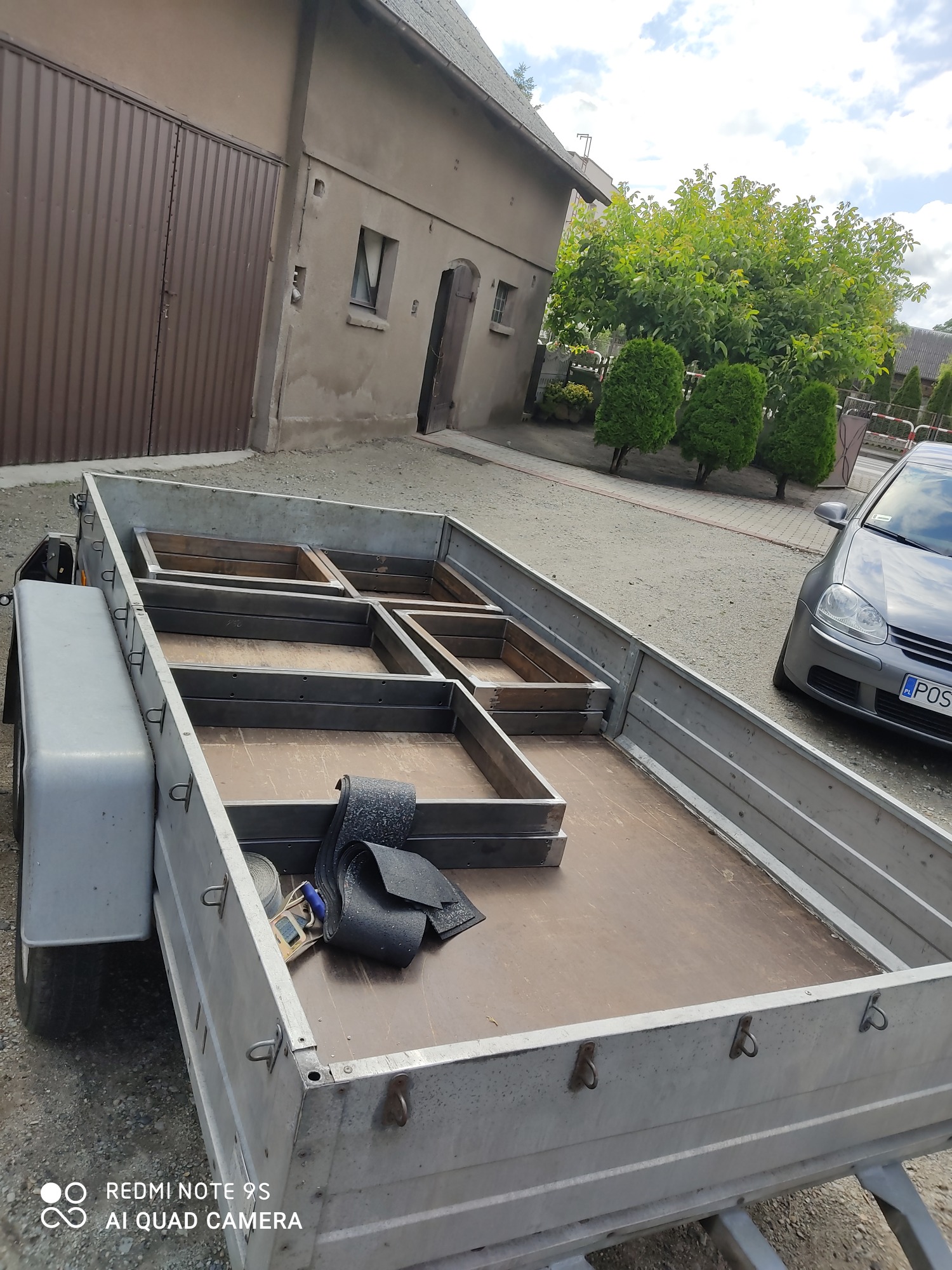 Przyczepa z metalową konstrukcją kratową wewnątrz, prawdopodobnie w trakcie budowy lub modyfikacji, z widocznymi narzędziami i pasami mocującymi, na tle budynku gospodarczego i zaparkowanego...