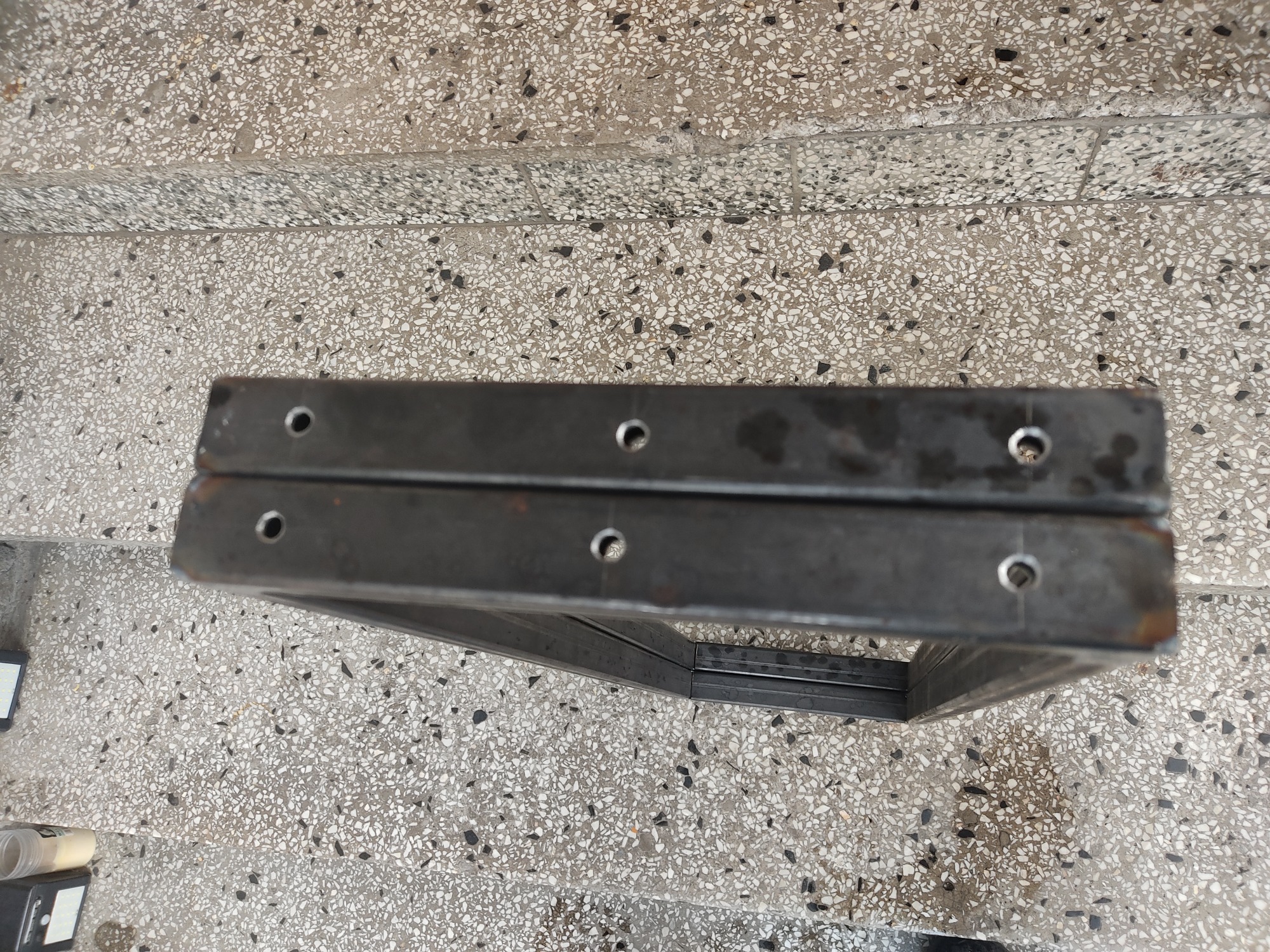 Dwa metalowe, prostokątne elementy z otworami, ułożone jeden na drugim na schodach o wzorzystej, kamiennej powierzchni.