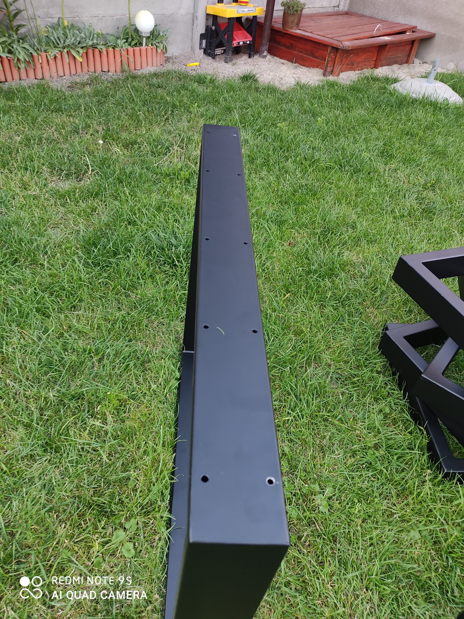 Czarne, metalowe elementy konstrukcyjne z otworami, leżące na trawie, prawdopodobnie przygotowane do montażu. Widoczne spawane łączenia.