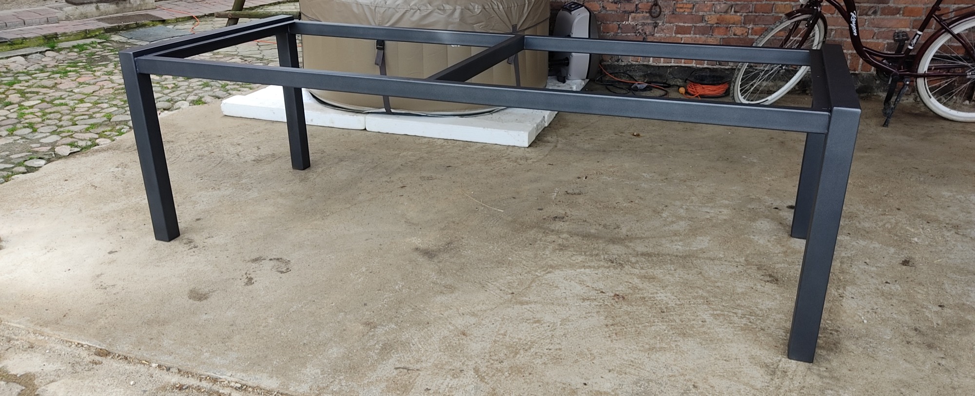 Czarna, metalowa rama stołu z kwadratowych profili, stojąca na betonowym podłożu, z widocznym w tle basenem ogrodowym i rowerem opartym o ceglany mur.