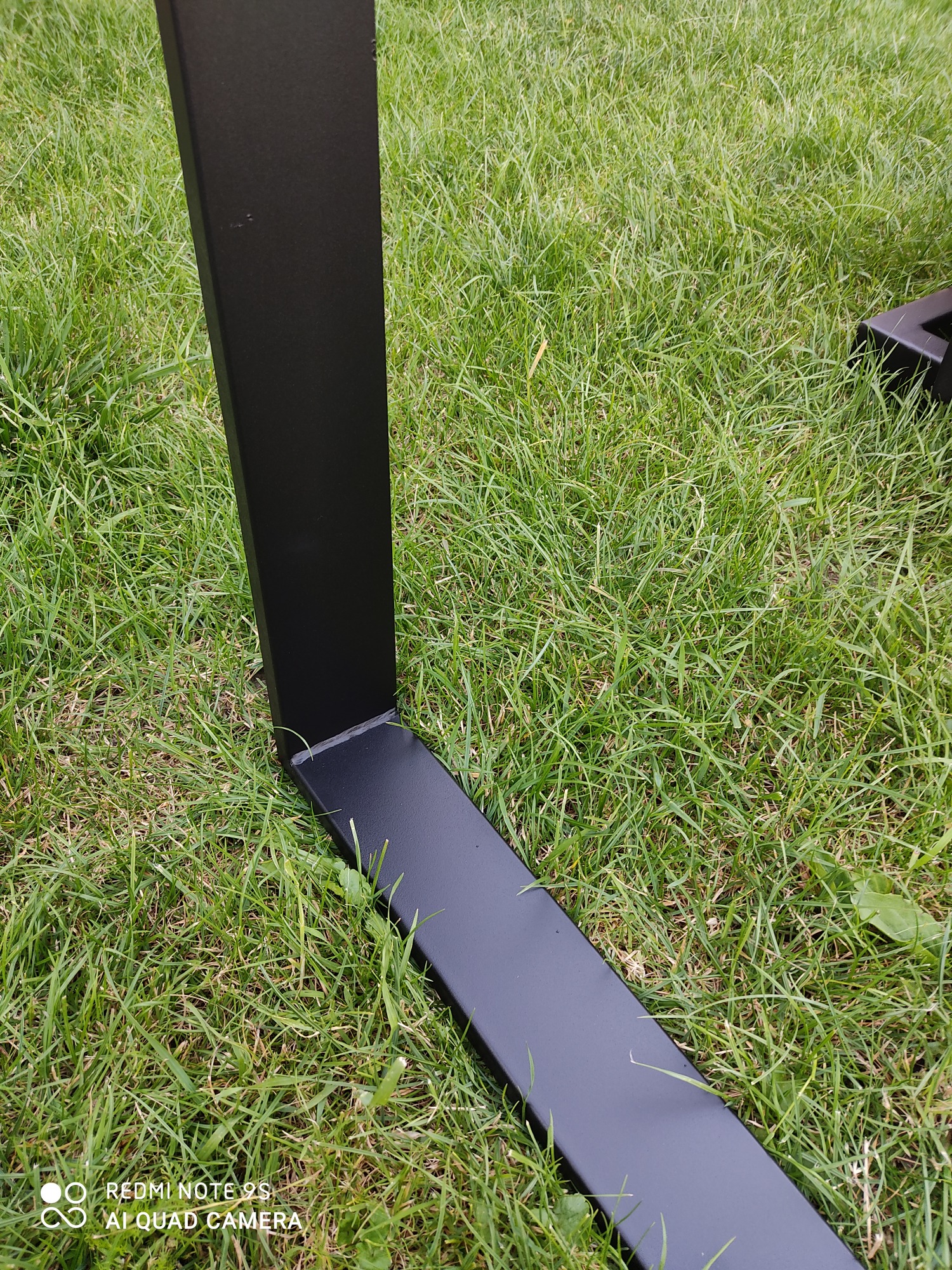 Czarne, metalowe nogi stołu spawane pod kątem prostym, leżące na trawie w słoneczny dzień.