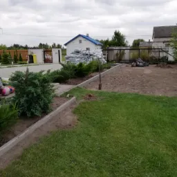 Przygotowany teren pod przyszły ogród z widocznymi obrzeżami, workami ziemi i narzędziem ogrodniczym. Część trawnika zachowana, dom w tle.