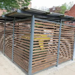 Zadaszona konstrukcja osłaniająca pojemniki na śmieci, z szarymi metalowymi słupkami i poziomymi drewnianymi listwami, na zewnątrz budynku z czerwonej cegły.
