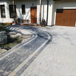 Nowo ułożony podjazd z kostki brukowej w dwóch odcieniach szarości, z dekoracyjnym łukiem prowadzącym do domu z brązowymi drzwiami i garażem.
