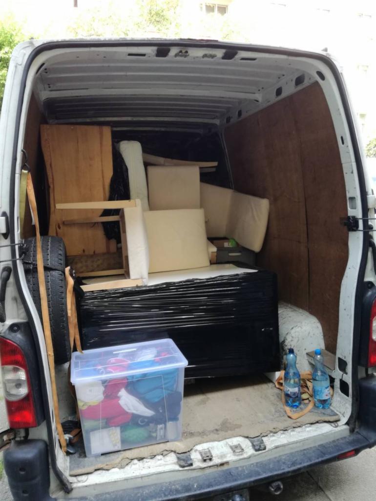 Wnętrze białego busa załadowane meblami, materacem owiniętym czarną folią, kartonami i oponą, przygotowane do transportu podczas przeprowadzki.