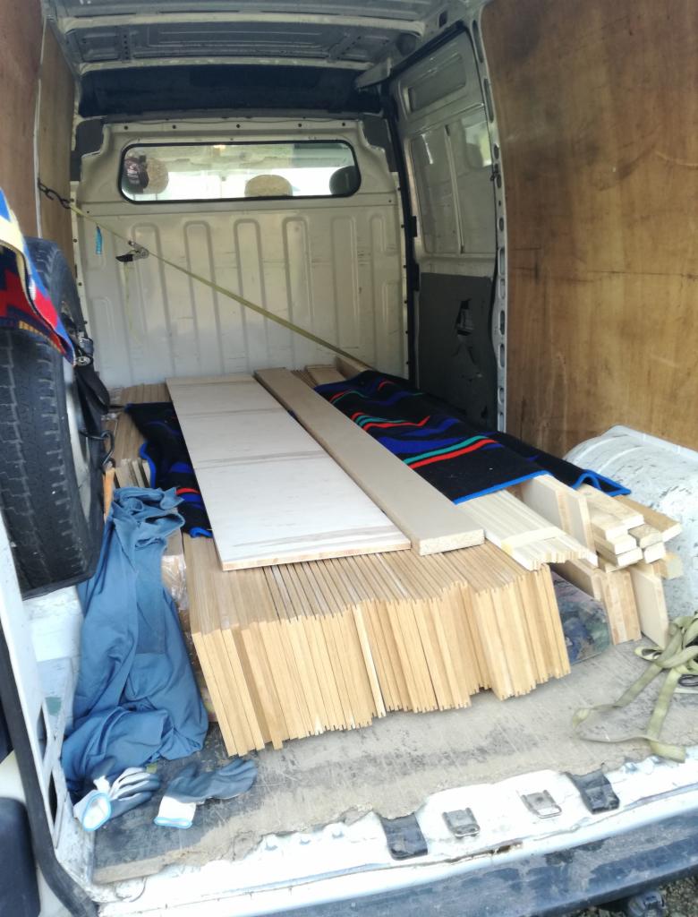 Wnętrze białego busa z załadowanym stosem drewnianych desek, przykrytych częściowo kocem w kolorowe paski, zabezpieczonych pasami transportowymi.