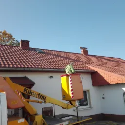 Pracownik w podnośniku koszowym marki Kiloutou, na tle domu z czerwoną dachówką, prawdopodobnie czyści rynnę.