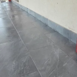 Szare, błyszczące płytki gresowe ułożone na zewnątrz budynku, z widoczną listwą przypodłogową z tego samego materiału.