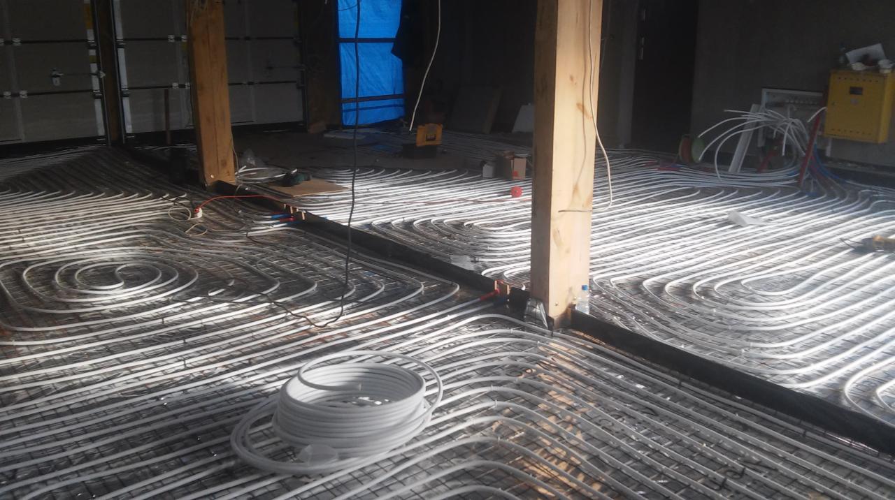 Montaż ogrzewania podłogowego: białe rury ułożone spiralnie na folii izolacyjnej w surowym wnętrzu, widoczne drewniane słupy konstrukcyjne i zwój rur.