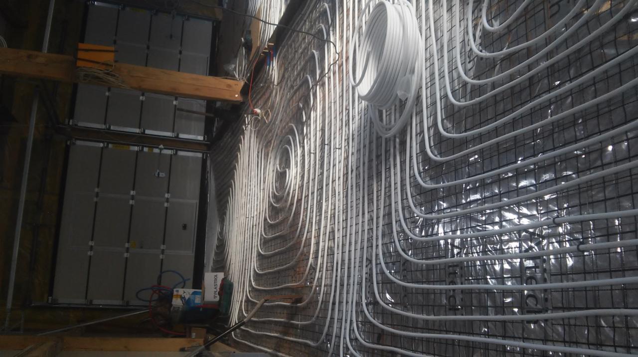 Montaż ogrzewania podłogowego ściennego: białe rurki układane spiralnie na metalowej siatce, widoczne materiały izolacyjne i konstrukcja drewniana.