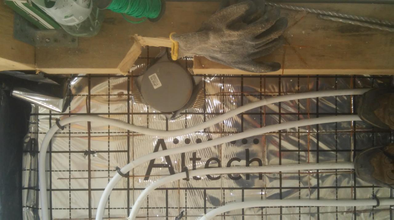 Montaż ogrzewania podłogowego: białe rury układane na folii refleksyjnej z siatką, widoczne narzędzia, rękawice robocze i fragment butów osoby wykonującej instalację.