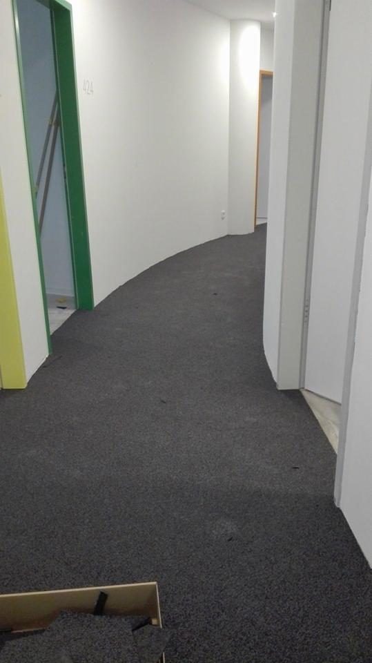 Korytarz biurowy z nową, ciemnoszarą wykładziną dywanową w trakcie układania, widoczny fragment opakowania wykładziny w rogu kadru, białe ściany i zielone obramowanie drzwi do pomieszczenia...