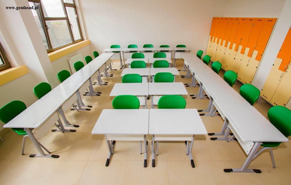Jasna sala lekcyjna z rzędami białych stolików i zielonych krzeseł, pomarańczowo-beżowe szafki w tle, widok z góry na ustawienie mebli.