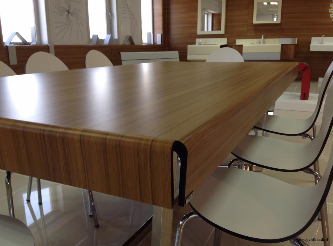 Stół z blatem imitującym drewno i metalowymi nogami, otoczony białymi krzesłami w showroomie we Wrocławiu, z widocznymi elementami wyposażenia łazienki w tle.