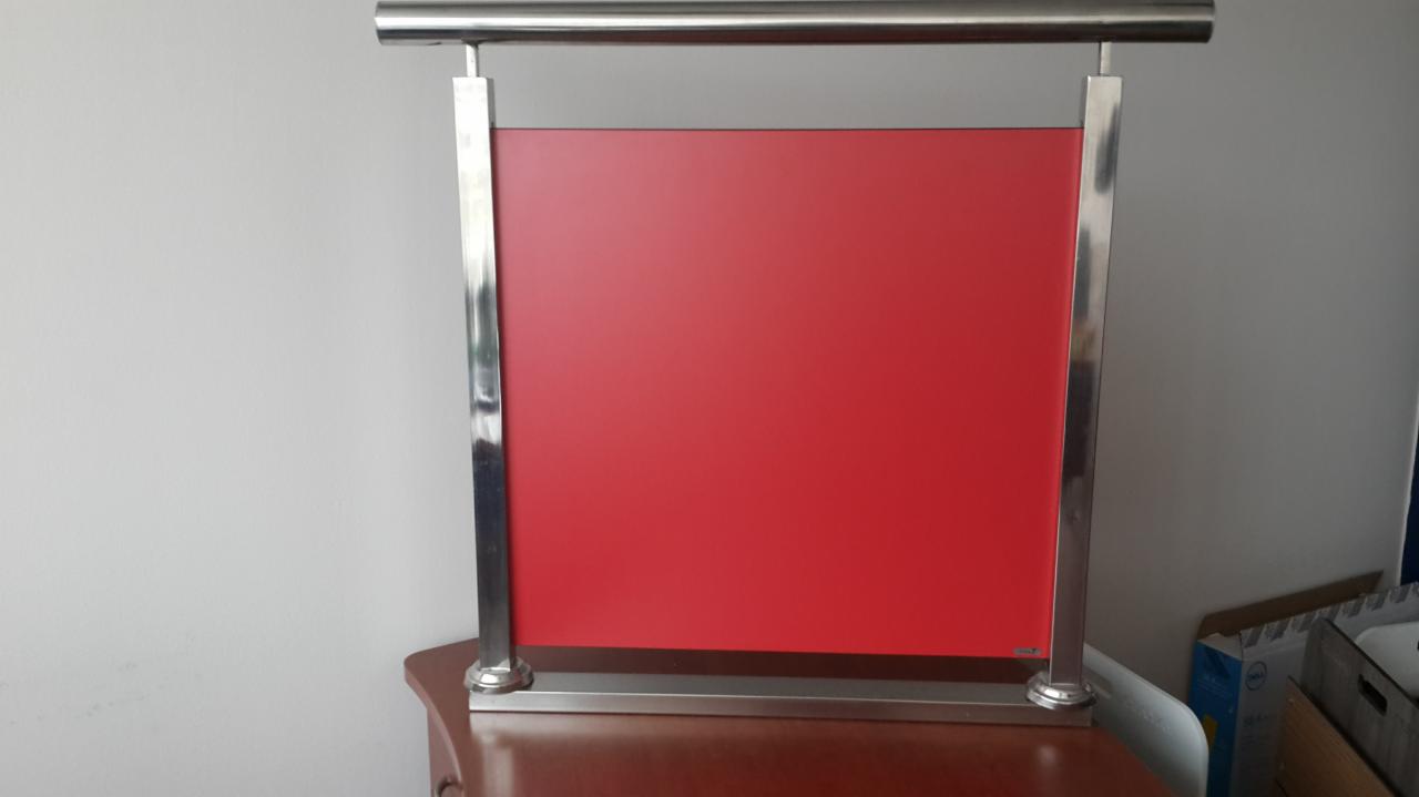 Czerwony panel zamontowany w metalowej ramie z elementami chromowanymi, umieszczony na drewnianym blacie, prawdopodobnie fragment ekspozycji meblowej.