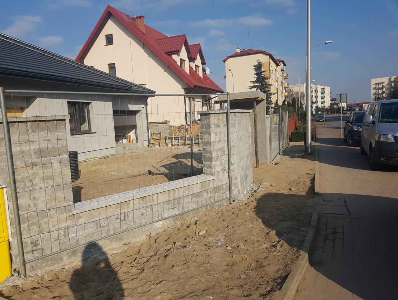 Wznoszona konstrukcja ogrodzenia z szarej kostki betonowej, zabezpieczona siatką, na tle budynków mieszkalnych i ulicy w słoneczny dzień.