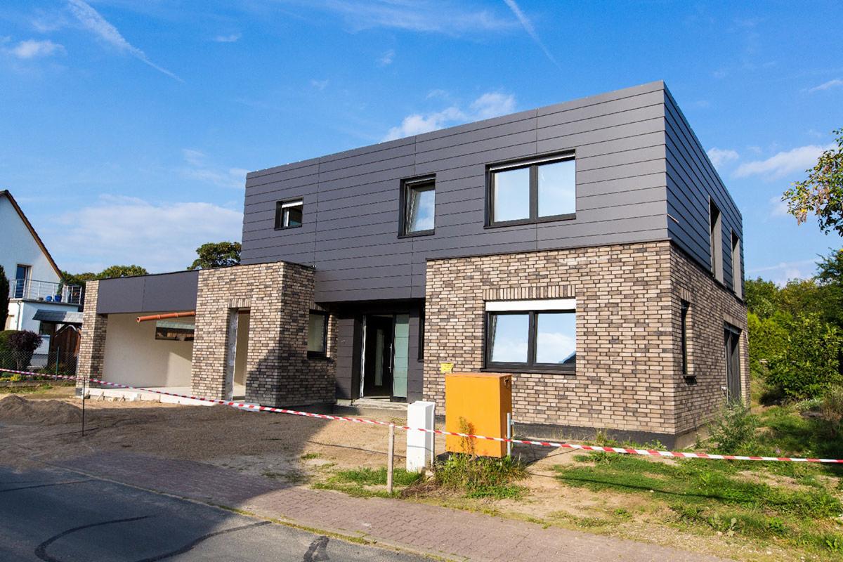 Dwukondygnacyjny dom z szarą elewacją i ceglaną fasadą, widoczny pomarańczowy licznik gazu, otoczony taśmą ostrzegawczą, na tle błękitnego nieba.