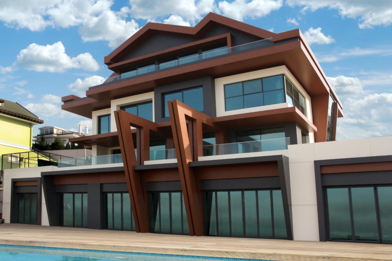 Nowoczesny, trzypiętrowy dom z elementami drewnianymi, balkonami z przezroczystymi balustradami i dużymi oknami, widziany z perspektywy basenu w słoneczny dzień.