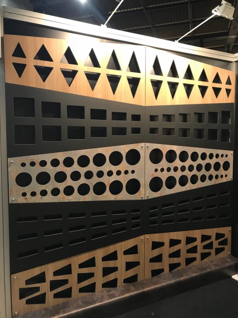 Panel dekoracyjny z wyciętymi wzorami: trójkąty, kwadraty, koła i prostokąty, ułożone warstwowo na drewnianym i metalowym tle.