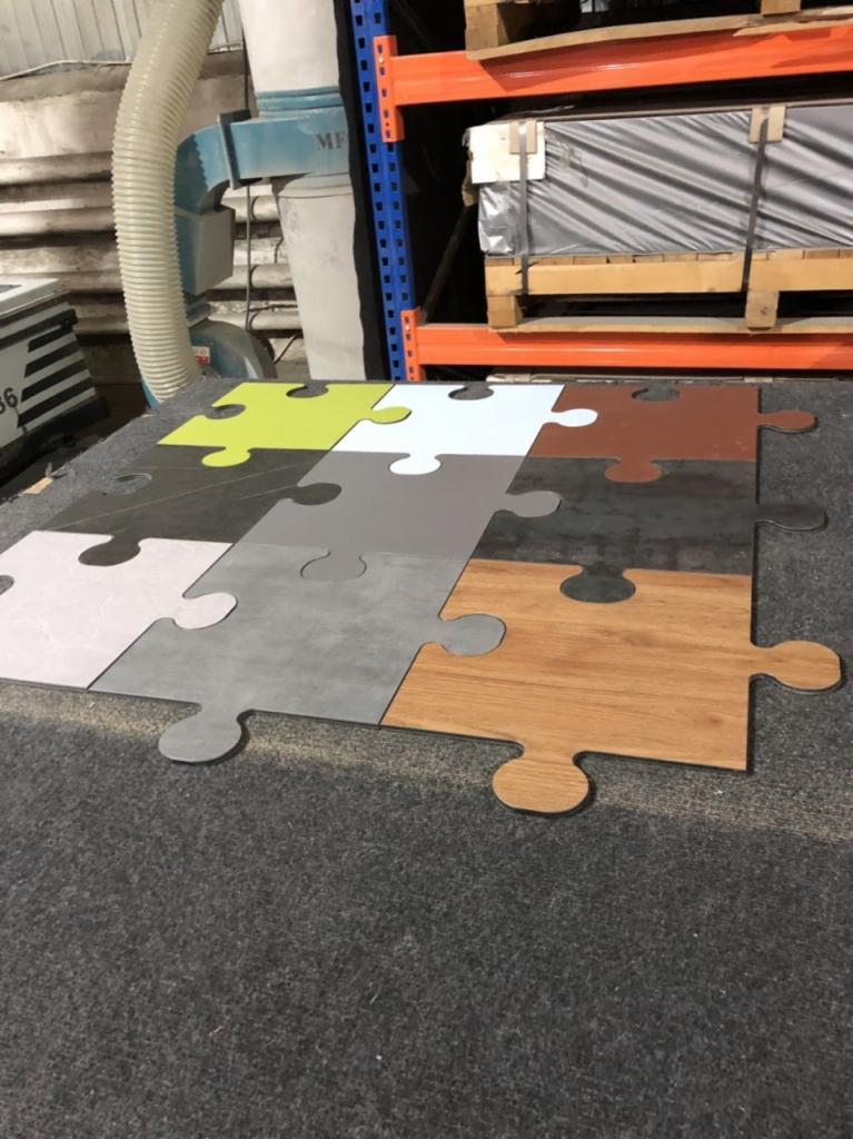 Puzzle z fragmentami różnych materiałów wykończeniowych: drewno, ceramika, beton, tworzące mozaikę kolorów i faktur na tle szarej wykładziny, z regałem magazynowym w tle.