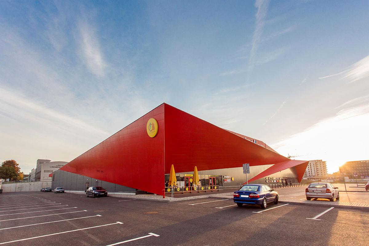 Nowoczesny budynek komercyjny z czerwoną elewacją i żółtym okrągłym logo, widok z parkingu z zaparkowanymi samochodami w słoneczny dzień.
