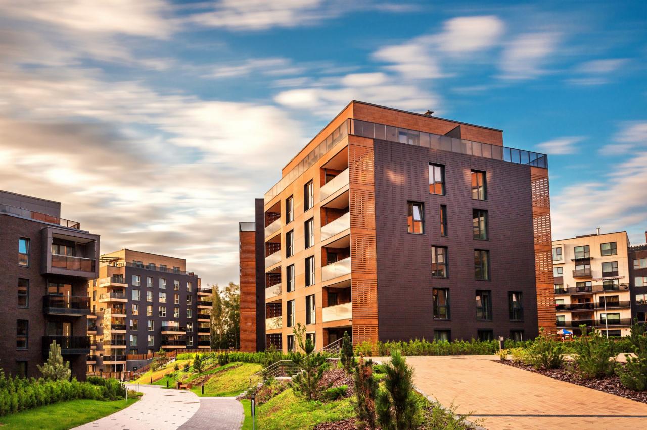 Nowoczesne budynki wielorodzinne z elewacją z ciemnej cegły i drewnianymi panelami, balkony z przeszklonymi balustradami, otoczone zielenią i ścieżkami spacerowymi, na tle błękitnego nieba z chmurami.