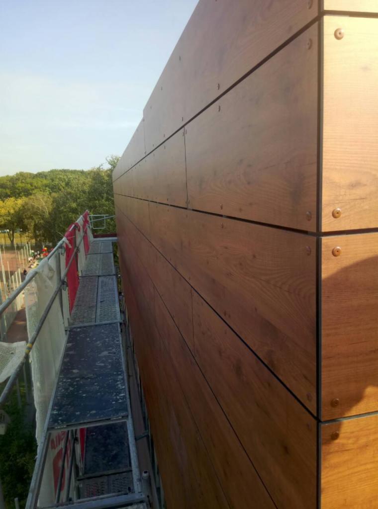 Fragment ściany budynku z okładziną imitującą drewno, widoczny ze stojącego obok rusztowania, z elementami mocującymi na rogach paneli.