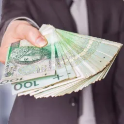 Dłoń w mankiecie marynarki trzyma plik banknotów 100 zł z portretem króla Władysława Jagiełły.