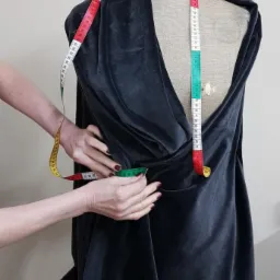 Krawcowa mierzy ciemny materiał na manekinie krawieckim za pomocą miarki krawieckiej. Widoczne dłonie i fragment materiału. Kadr od frontu, oświetlenie naturalne.